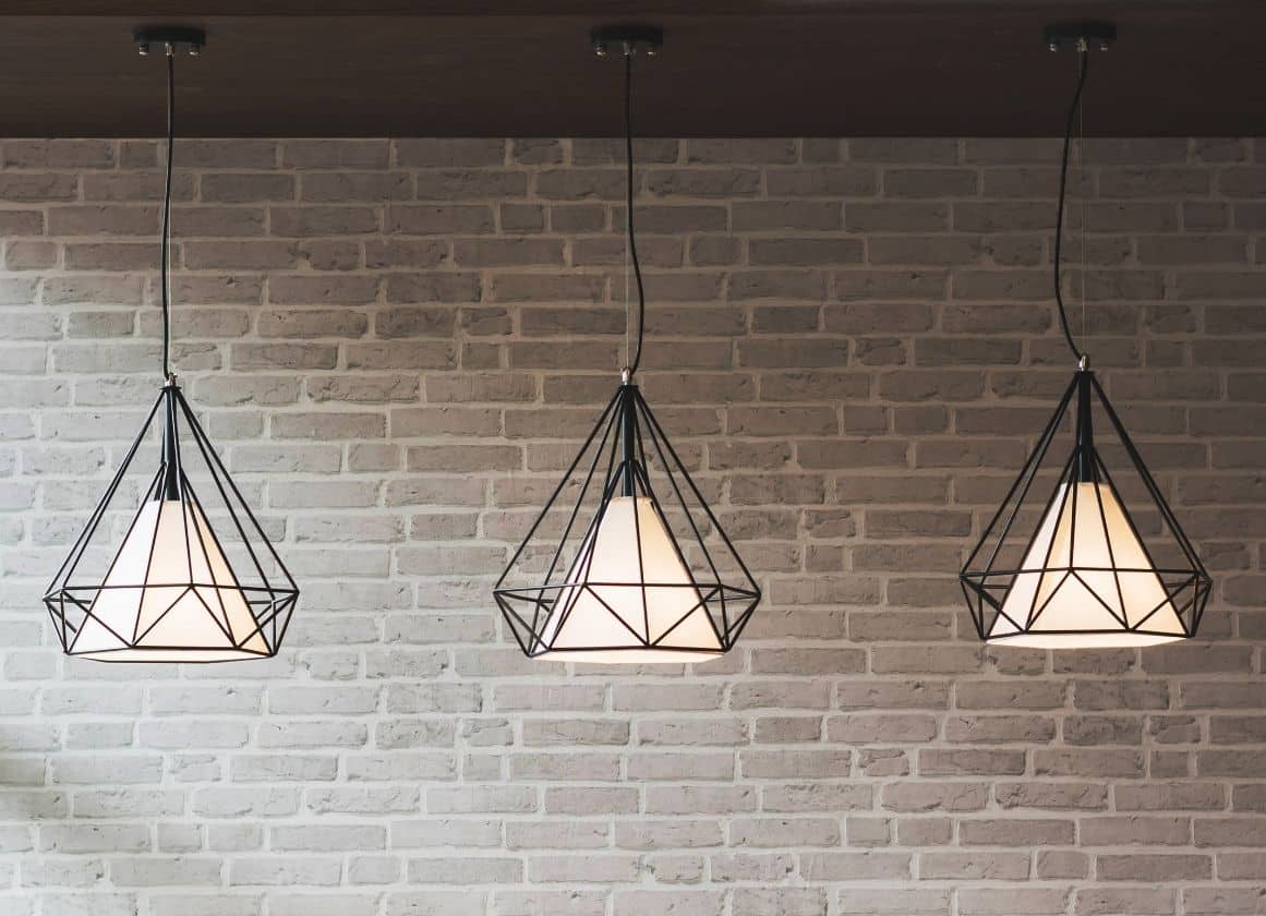 Trois suspensions lumineuses design installées au plafond devant un mur en briques claires dans un intérieur contemporain.